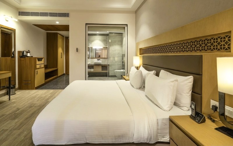 Classic Room — king bed, wooden accents and glass-walled bath at Excel Hotels & Resorts, Jim Corbett